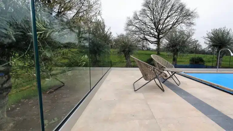 Flat poolside patio with a frameless glass balustrade railing looking out to a large open grass field with a distance tree line