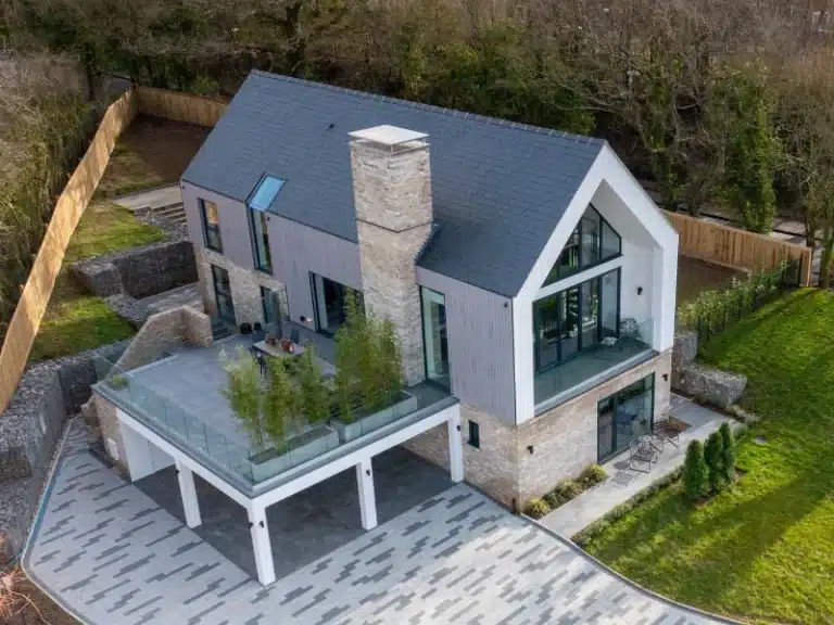 arial view of a large house surrounded by a wood fences and trees. the house has a raised stone patio with frameless glass balustrades and a set of clean garden furniture