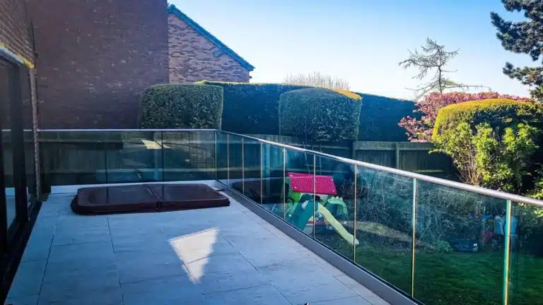 pale stone patio with a glass balustrade railing in the shade overlooking a garden with bushes and children's toys