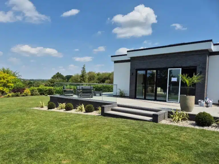 A raised garden patio with a glass balustrade railing overlooking a garden with a gravel strip and well trimmed bushes