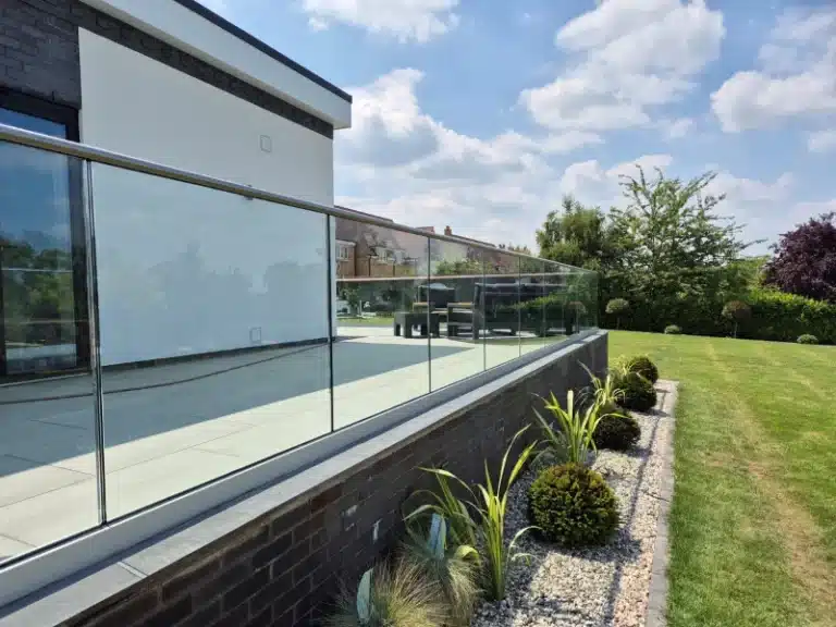 A raised garden patio with a glass balustrade railing overlooking a garden with a gravel strip and well trimmed bushes