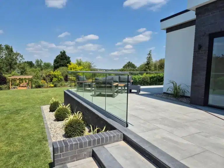 A raised garden patio with a glass balustrade railing overlooking a garden with a gravel strip and well trimmed bushes