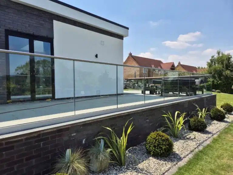 A raised garden patio with a glass balustrade railing overlooking a garden with a gravel strip and well trimmed bushes