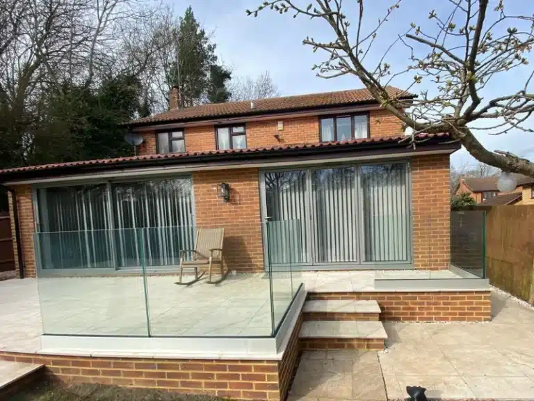 Pale grey stone tile patio outside a red brick house with frameless glass balustrades and a wooden rocking chair
