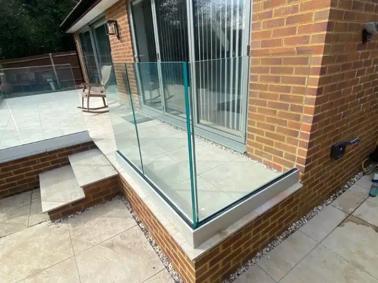 Pale grey stone tile patio outside a red brick house with frameless glass balustrades and a wooden rocking chair