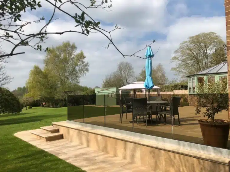 pale stone tile patio with wicker garden furniture and a frameless black stained glass balustrade looking out to a long garden with a lime green shed
