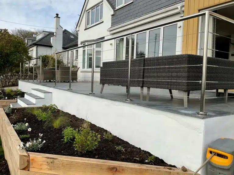 large clean modern home with a grey slate tile patio and thatched wood outdoor furniture separated from plank bordered flower beds by metal wire balustrades