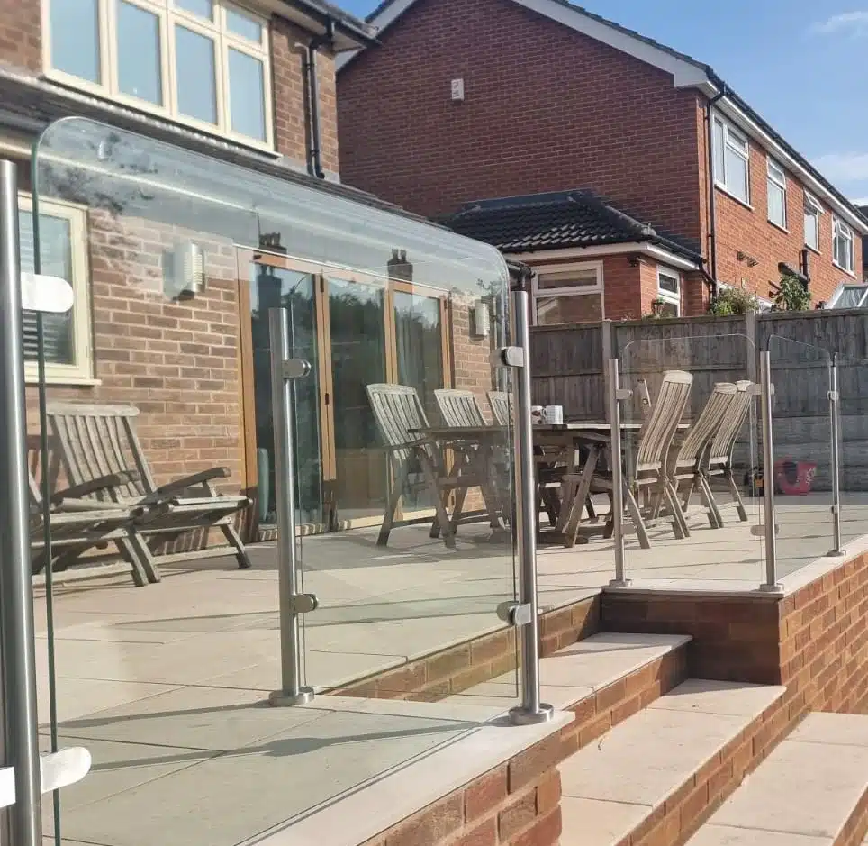 Patio with wooden garden furniture and a rounded glass balustrade fence outside a clean house