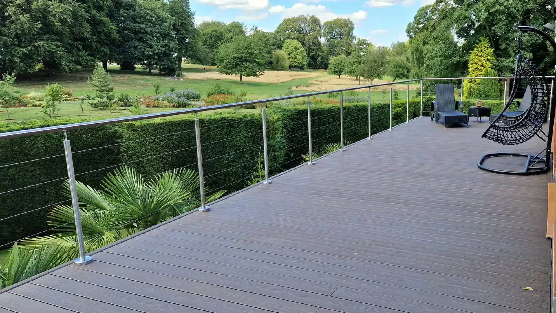Dark wooden patio with wire balustrade facing green field filled with tall trees